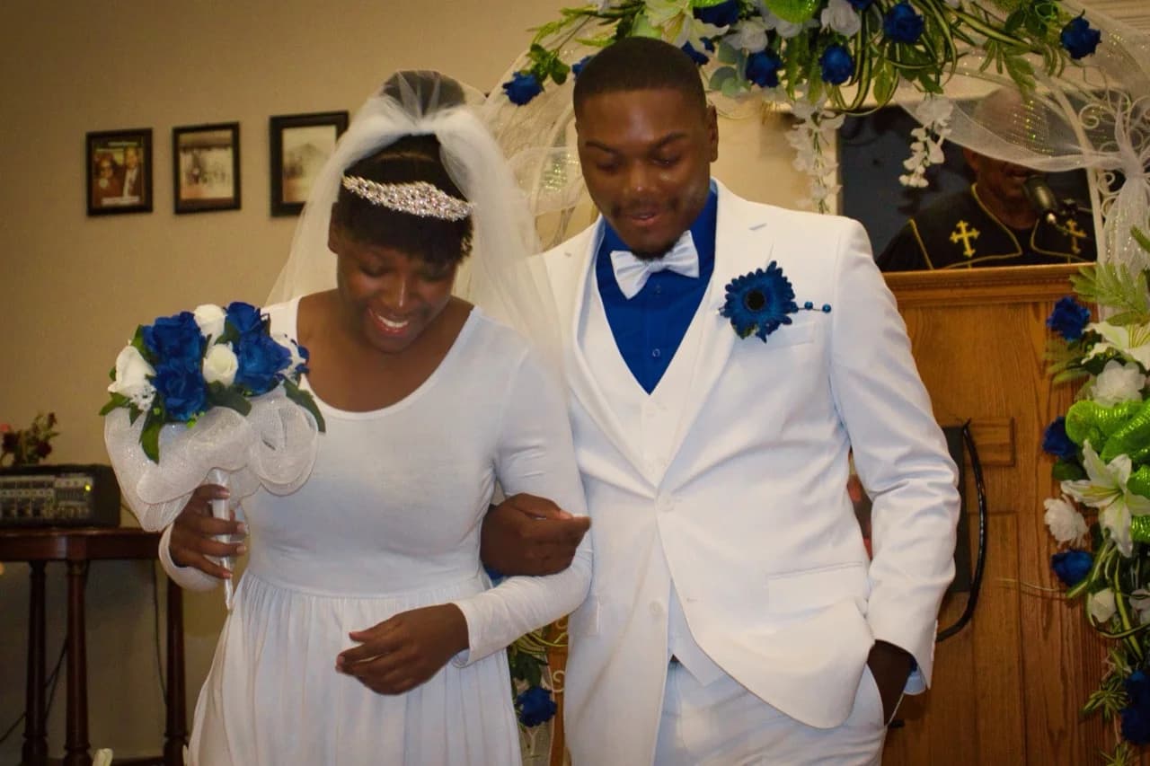 Sydney and Melvin smiling, arm in arm on their wedding day, dressed in white and blue